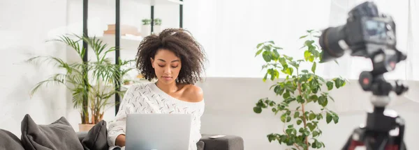 Prise de vue panoramique d'un blogueur afro-américain utilisant un ordinateur portable près d'un appareil photo numérique — Photo de stock