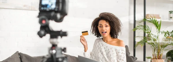 Panoramic shot of surprised african american video blogger holding credit card near digital camera — Stock Photo