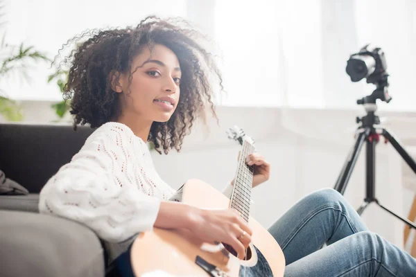 Joven y rizado afroamericano chica en frenos celebración de guitarra acústica cerca de la cámara digital - foto de stock