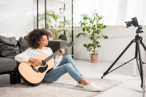 Messa a fuoco selettiva di giovane e riccia ragazza africana americana che suona la chitarra acustica vicino alla fotocamera digitale in soggiorno — Foto stock