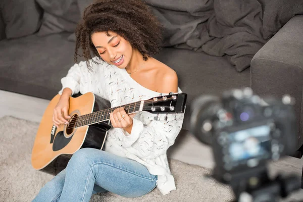 Messa a fuoco selettiva di felice ragazza afroamericana in bretelle suonare la chitarra acustica vicino alla fotocamera digitale in soggiorno — Foto stock