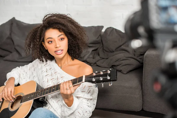 Селективное внимание молодой африканской девушки в брекетах, играющей на акустической гитаре и смотрящей на цифровую камеру в гостиной — стоковое фото