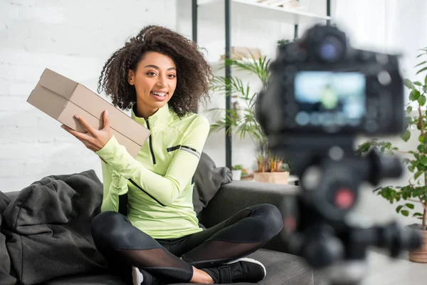 Selective focus of sportive african american influencer holding carton box near digital camera — Stock Photo