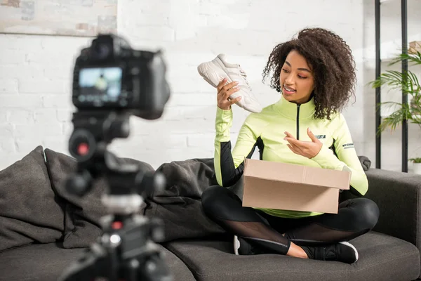 Foco seletivo do esportivo afro-americano influenciador em aparelhos segurando novo tênis perto da caixa e câmera digital — Fotografia de Stock