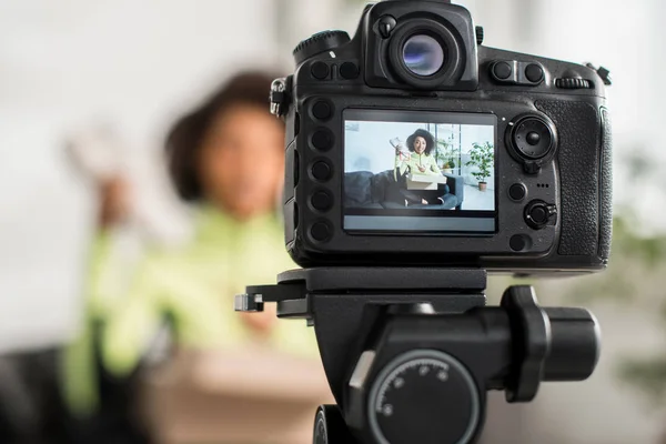 Selective focus of  digital camera  with sportive african american influencer holding new sneaker near box on display — Stock Photo