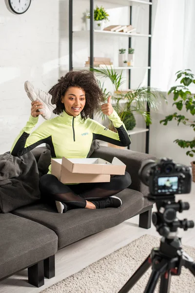 Selective focus of cheerful african american influencer in braces showing thumb up and holding new sneaker near digital camera — Stock Photo