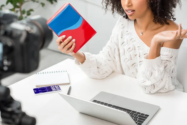 Vista ritagliata di influencer afro-americano in bretelle che tengono scatola colorata vicino smartphone con app medica sullo schermo e fotocamera digitale — Foto stock