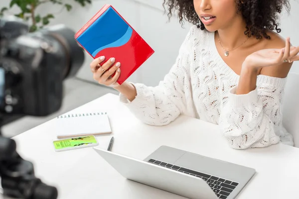 Vista recortada do afro-americano influenciador em aparelho segurando caixa colorida perto de smartphone com melhor aplicativo de compras na tela e câmera digital — Fotografia de Stock