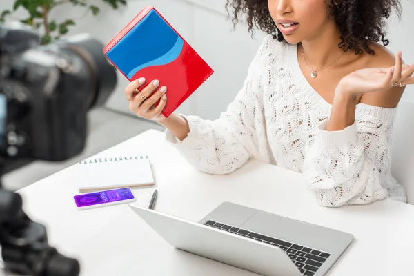 Vista recortada do afro-americano influenciador em aparelho segurando caixa colorida perto de smartphone com aplicativo de compras e câmera digital — Fotografia de Stock