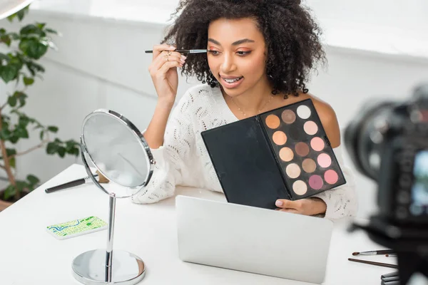 Foyer sélectif de joyeux blogueur vidéo afro-américain dans les bretelles appliquant ombre à paupières près du smartphone avec carte en ligne et appareil photo numérique — Photo de stock