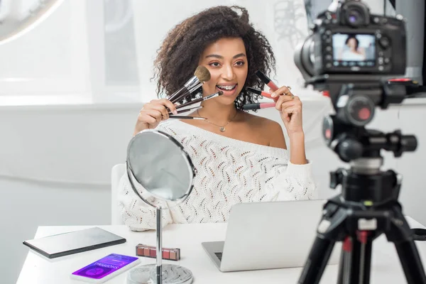 Selective focus of happy african american influencer in braces holding lip glosses and cosmetic brushes near smartphone with shopping app and digital camera — Stock Photo