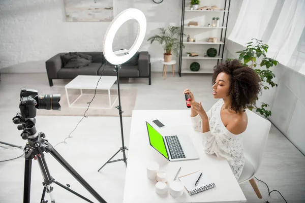 Influencer afroamericano con piastra per capelli vicino alla fotocamera digitale e laptop con schermo verde — Foto stock