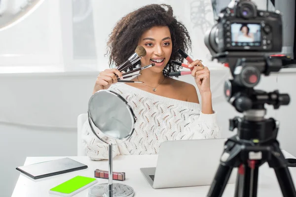 Selective focus of happy african american influencer in braces holding lip glosses and cosmetic brushes near smartphone with green screen and digital camera — Stock Photo