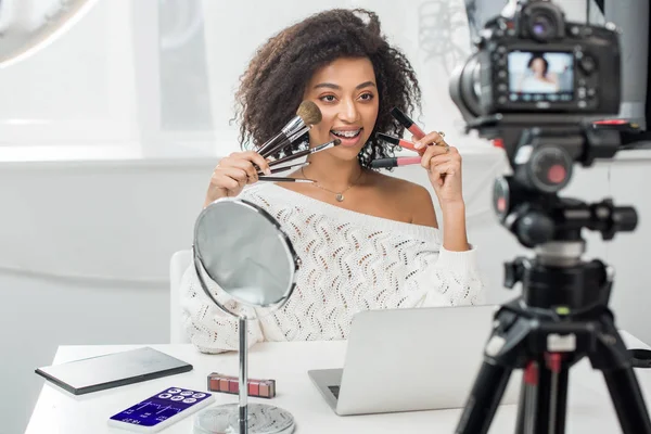 Messa a fuoco selettiva di felice influencer afro-americano in bretelle con lucidalabbra e pennelli cosmetici vicino smartphone con tracker medico e fotocamera digitale — Foto stock