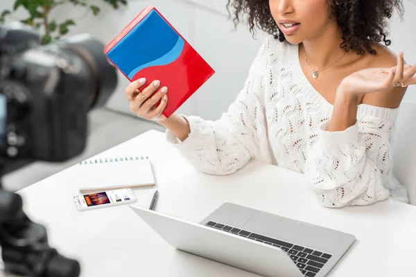 KYIV, UCRANIA - 10 DE DICIEMBRE DE 2019: vista recortada de una chica afroamericana con tirantes sosteniendo una caja de colores cerca de un teléfono inteligente con aplicación soundcloud en la pantalla y cámara digital - foto de stock