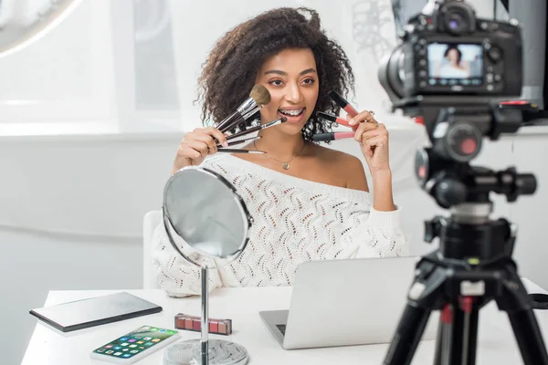 KYIV, UKRAINE - DECEMBER 10, 2019: selective focus of happy african american girl in braces holding lip glosses and cosmetic brushes near iphone — Stock Photo