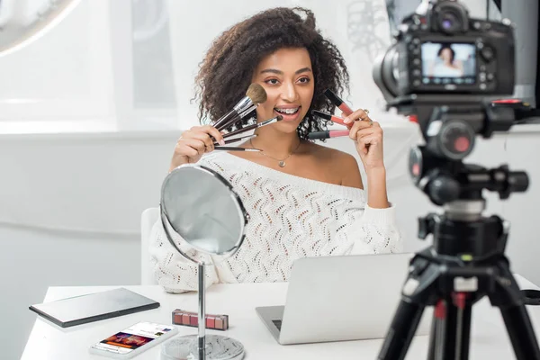 KYIV, UKRAINE - DECEMBER 10, 2019: selective focus of african american woman in braces holding lip glosses and cosmetic brushes near smartphone with soundcloud app — Stock Photo