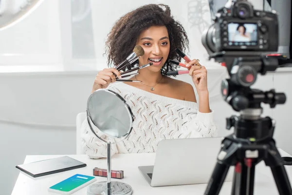KYIV, UKRAINE - DECEMBER 10, 2019: selective focus of african american girl in braces holding lip glosses and cosmetic brushes near smartphone with twitter app — Stock Photo