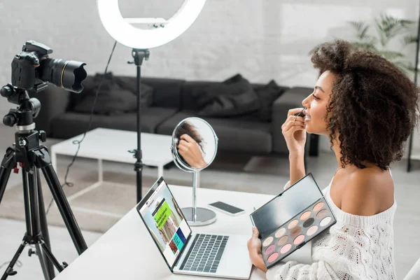 KYIV, UKRAINE - DEZEMBRO 10, 2019: visão lateral do afro-americano influenciador segurando paleta de sombra de olhos perto do laptop com site bbc e câmera digital — Fotografia de Stock