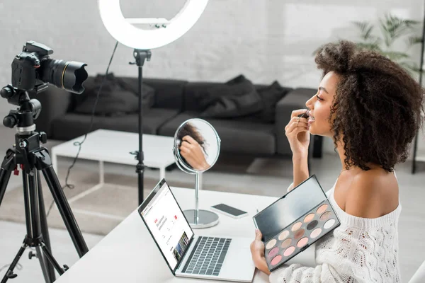 КИЕВ, УКРАИНА - 10 ДЕКАБРЯ 2019 года: вид сбоку африканского американского влияния, держащего палитру теней для век рядом с ноутбуком с веб-сайтом airbnb и цифровой камерой — стоковое фото