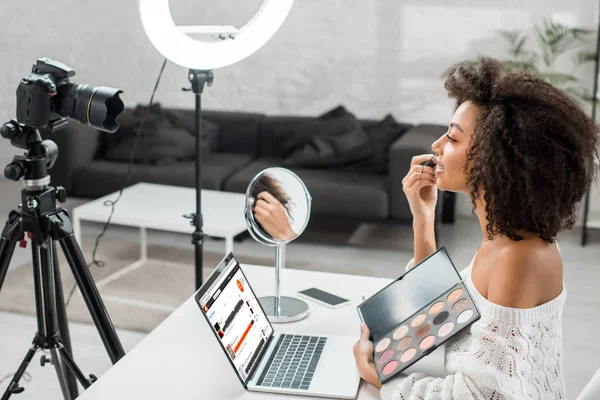 KYIV, UKRAINE - DEZEMBRO 10, 2019: visão lateral do afro-americano influenciador segurando paleta de sombra de olhos perto do laptop com site soundcloud e câmera digital — Fotografia de Stock