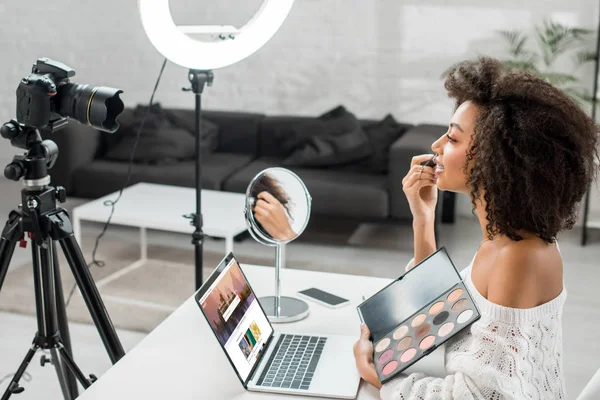 KYIV, UKRAINE - DEZEMBRO 10, 2019: visão lateral do afro-americano influenciador segurando paleta de sombra de olhos perto do laptop com site shutterstock e câmera digital — Fotografia de Stock