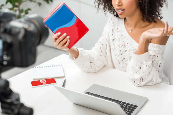 KYIV, UKRAINE - DECEMBER 10, 2019: cropped view of african american influencer in braces holding colorful box near smartphone with youtube app on screen and digital camera — Stock Photo