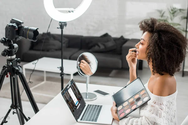 KYIV, UKRAINE - DEZEMBRO 10, 2019: visão lateral do afro-americano influenciador segurando paleta de sombra de olhos perto do laptop com site linkedin e câmera digital — Fotografia de Stock