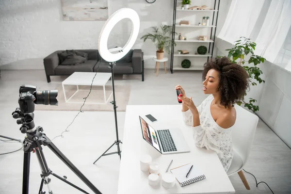 KYIV, UCRANIA - 10 DE DICIEMBRE DE 2019: Afroamericano influencer holding hair straightener near digital camera and laptop with shutterstock website - foto de stock