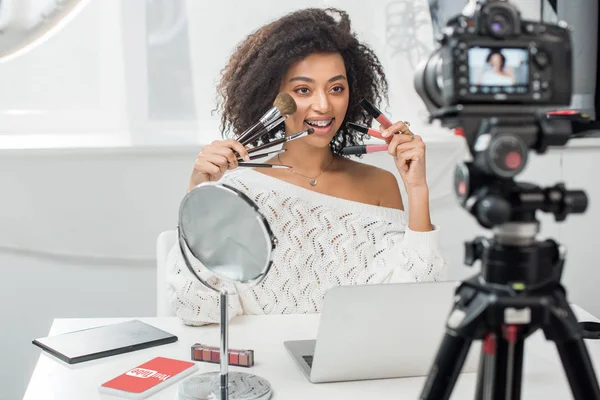 KYIV, UKRAINE - DECEMBER 10, 2019: selective focus of african american woman in braces holding lip glosses and cosmetic brushes near smartphone with youtube app — Stock Photo