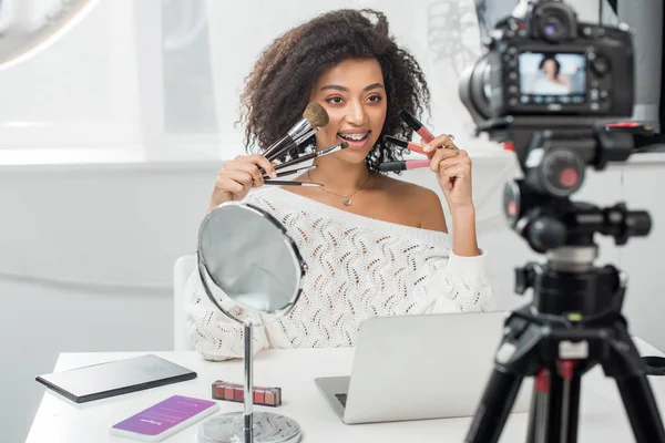 KYIV, UKRAINE - DECEMBER 10, 2019: selective focus of african american woman in braces holding lip glosses and cosmetic brushes near smartphone with instagram app — Stock Photo