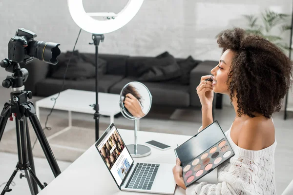 KYIV, UKRAINE - DECEMBER 10, 2019: side view of african american influencer holding eye shadow palette near laptop with shutterstock website and digital camera — Stock Photo