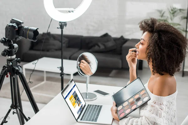 KYIV, UKRAINE - DEZEMBRO 10, 2019: visão lateral do afro-americano influenciador segurando paleta de sombra de olhos perto do laptop com site de reserva e câmera digital — Fotografia de Stock