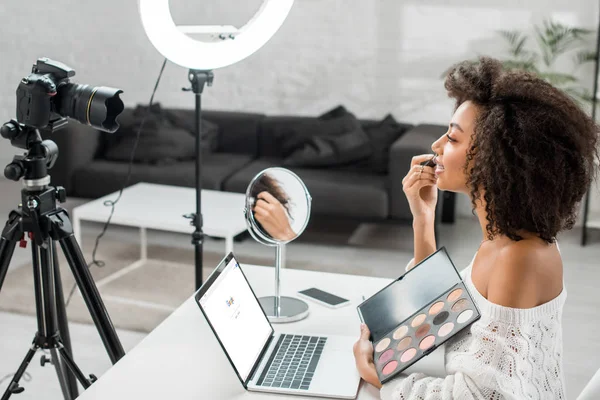KYIV, UKRAINE - DECEMBER 10, 2019: side view of african american influencer holding eye shadow palette near laptop with google website and digital camera — Stock Photo