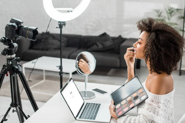 KYIV, UKRAINE - DEZEMBRO 10, 2019: visão lateral do afro-americano influenciador segurando paleta de sombra olho perto do laptop com o site do google e câmera digital — Fotografia de Stock