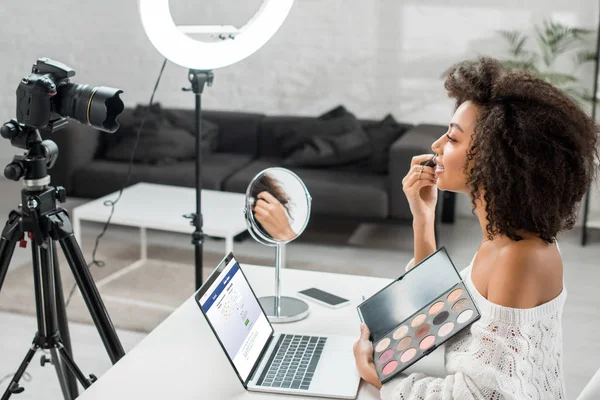 KYIV, UKRAINE - DEZEMBRO 10, 2019: visão lateral do afro-americano influenciador segurando paleta de sombra de olhos perto do laptop com site do Facebook e câmera digital — Fotografia de Stock