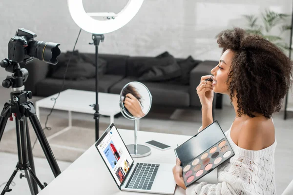KYIV, UKRAINE - DECEMBER 10, 2019: side view of african american influencer holding eye shadow palette near laptop with ebay website and digital camera — Stock Photo