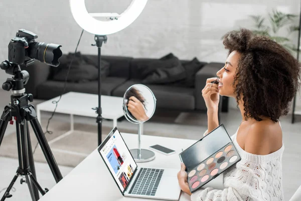 KYIV, UKRAINE - DEZEMBRO 10, 2019: visão lateral do afro-americano influenciador segurando paleta de sombra de olhos perto do laptop com site do ebay e câmera digital — Fotografia de Stock