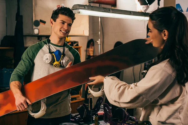 Woman giving snowboard to smiling worker in repair shop — Stock Photo
