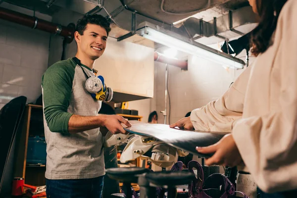 Cropped view of woman giving snowboard to smiling worker in repair shop — Stock Photo