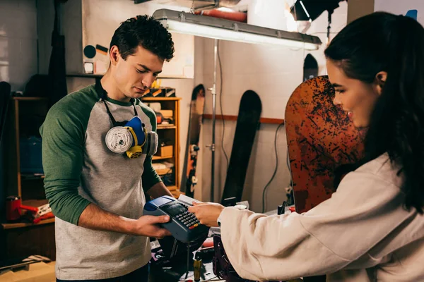 Worker holding terminal and smiling woman paying with credit card and holding snowboard in repair shop — Stock Photo