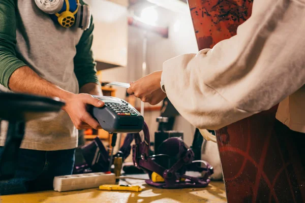 Cropped view of worker holding terminal and woman paying with credit card and holding snowboard in repair shop — Stock Photo