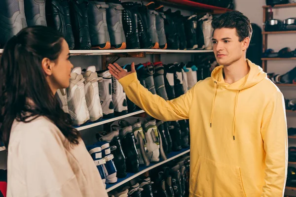 Trabajador señalando con la mano las botas de esquí y mirando al cliente en el taller de reparación - foto de stock