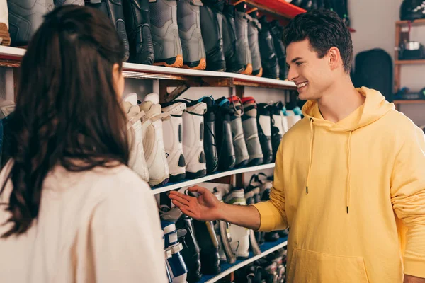 Enfoque selectivo del trabajador sonriente apuntando con la mano a las botas de esquí y el cliente mirándolo en el taller de reparación - foto de stock