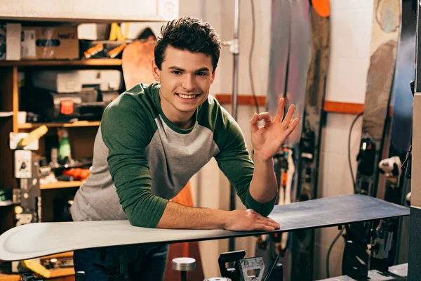 Smiling worker showing ok and standing near snowboard in repair shop — Stock Photo