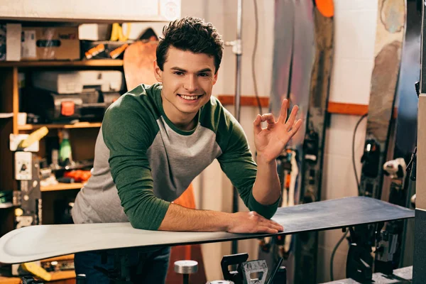 Sonriente trabajador mostrando ok y de pie cerca de snowboard en taller de reparación - foto de stock