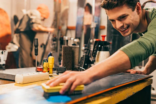 Smiling worker using brush on snowboard in repair shop — Stock Photo