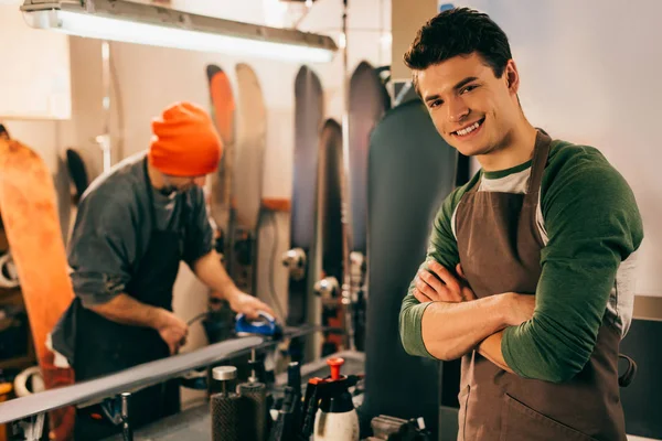 Messa a fuoco selettiva del lavoratore sorridente con le braccia incrociate e del suo collega che utilizza il ferro di cera in officina — Foto stock
