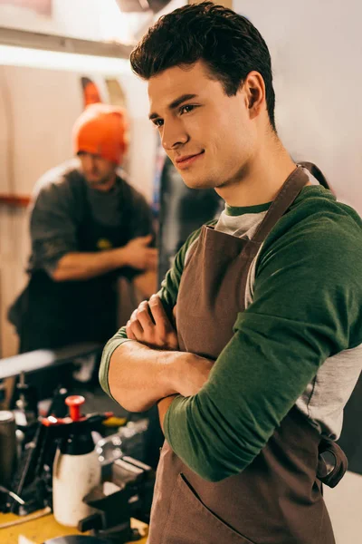 Selective focus of smiling worker with crossed arms and his colleague on background in repair shop — Stock Photo