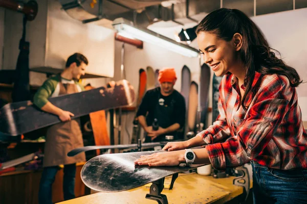 Focus selettivo del lavoratore sorridente che ripara snowboard e colleghi sullo sfondo — Foto stock